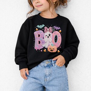 Puede incluir: Una sudadera negra de manga larga con un estampado gr&aacute;fico de la palabra BOO en rosa y morado pastel. El dise&ntilde;o incluye un fantasma gui&ntilde;ando un ojo con un lazo de lunares rosa, una calabaza naranja sonriente, murci&eacute;lagos y ma&iacute;z dulce.
