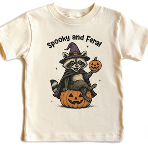Peut inclure: T-shirt cr&egrave;me avec un motif sur le th&egrave;me d'Halloween. Le dessin repr&eacute;sente un raton laveur portant un chapeau de sorci&egrave;re et une cape, assis sur une citrouille sculpt&eacute;e et tenant une lanterne. Le texte au-dessus du raton laveur dit "Spooky and Feral".