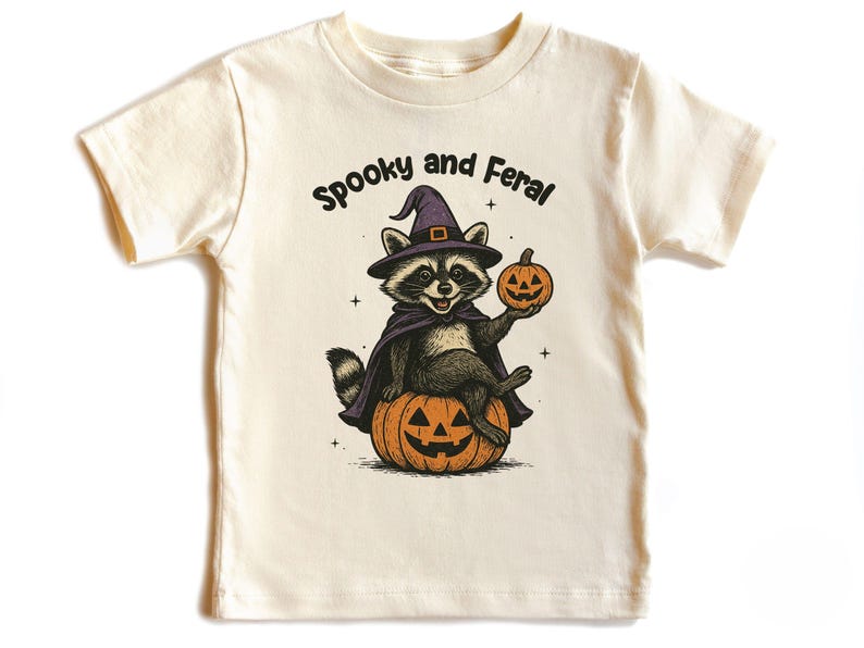 Peut inclure: T-shirt cr&egrave;me avec un motif sur le th&egrave;me d'Halloween. Le dessin repr&eacute;sente un raton laveur portant un chapeau de sorci&egrave;re et une cape, assis sur une citrouille sculpt&eacute;e et tenant une lanterne. Le texte au-dessus du raton laveur dit "Spooky and Feral".