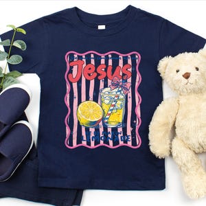 May include: Navy blue t-shirt with a retro graphic that says "Jesus Loves Lemonade". The graphic features a lemon, a glass of lemonade with a straw, and pink and blue stripes. A pair of navy blue shoes and a teddy bear are also in the image.