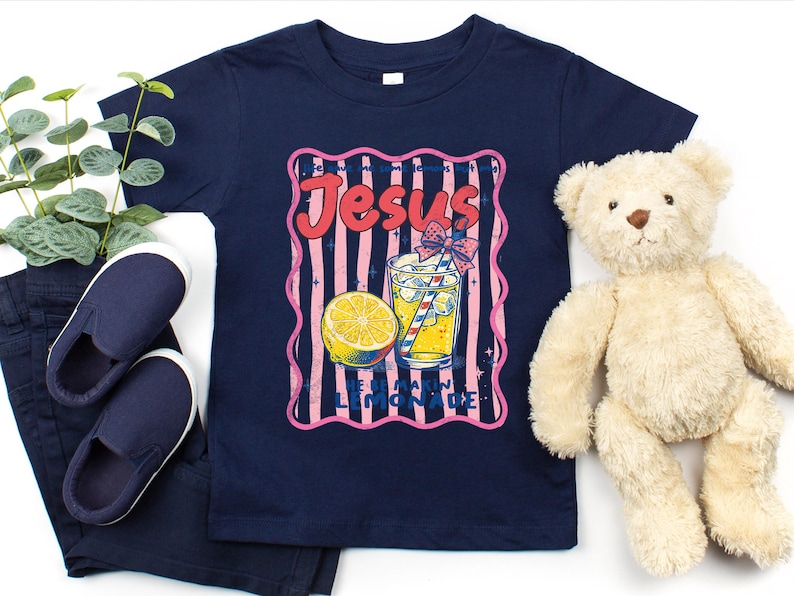 May include: Navy blue t-shirt with a retro graphic that says "Jesus Loves Lemonade". The graphic features a lemon, a glass of lemonade with a straw, and pink and blue stripes. A pair of navy blue shoes and a teddy bear are also in the image.