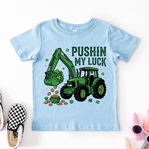 May include: Light blue toddler t-shirt with a green tractor graphic. The tractor has a bucket filled with shamrocks and is scooping up gold coins. Text reads "Pushin' My Luck". Black and white checkered slip-on shoes are in the image.