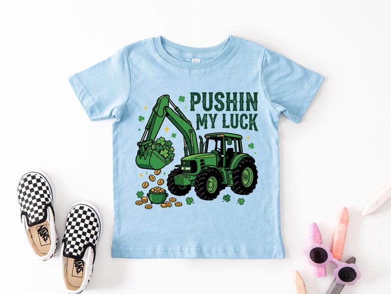 May include: Light blue toddler t-shirt with a green tractor graphic. The tractor has a bucket filled with shamrocks and is scooping up gold coins. Text reads "Pushin' My Luck". Black and white checkered slip-on shoes are in the image.