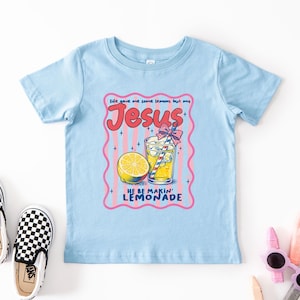 May include: A light blue t-shirt with a graphic that says "Life gave me some lemons but my Jesus, He be makin' lemonade." The graphic includes a lemon, a glass of lemonade, and a pink and red border.