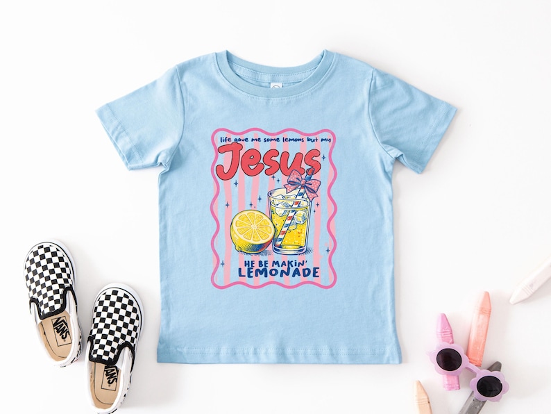 May include: A light blue t-shirt with a graphic that says "Life gave me some lemons but my Jesus, He be makin' lemonade." The graphic includes a lemon, a glass of lemonade, and a pink and red border.