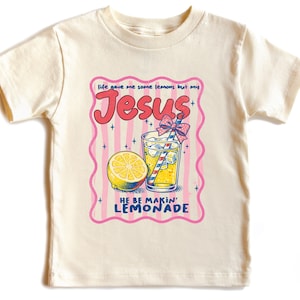 May include: Cream-colored t-shirt featuring a graphic of a lemon, a glass of lemonade with a straw, and the words "Jesus" and "He Be Makin' Lemonade" in a retro font. The design is framed by a pink border with vertical stripes.