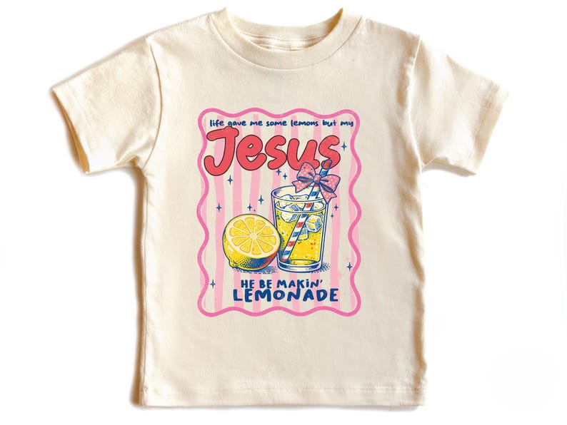 May include: Cream-colored t-shirt featuring a graphic of a lemon, a glass of lemonade with a straw, and the words "Jesus" and "He Be Makin' Lemonade" in a retro font. The design is framed by a pink border with vertical stripes.