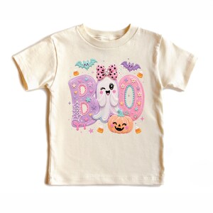 Puede incluir: Una camiseta de manga corta color crema con un estampado gr&aacute;fico de la palabra BOO en colores pastel. El dise&ntilde;o incluye un fantasma gui&ntilde;ando un ojo con un lazo rosa de lunares, una calabaza naranja sonriente, murci&eacute;lagos y telara&ntilde;as.