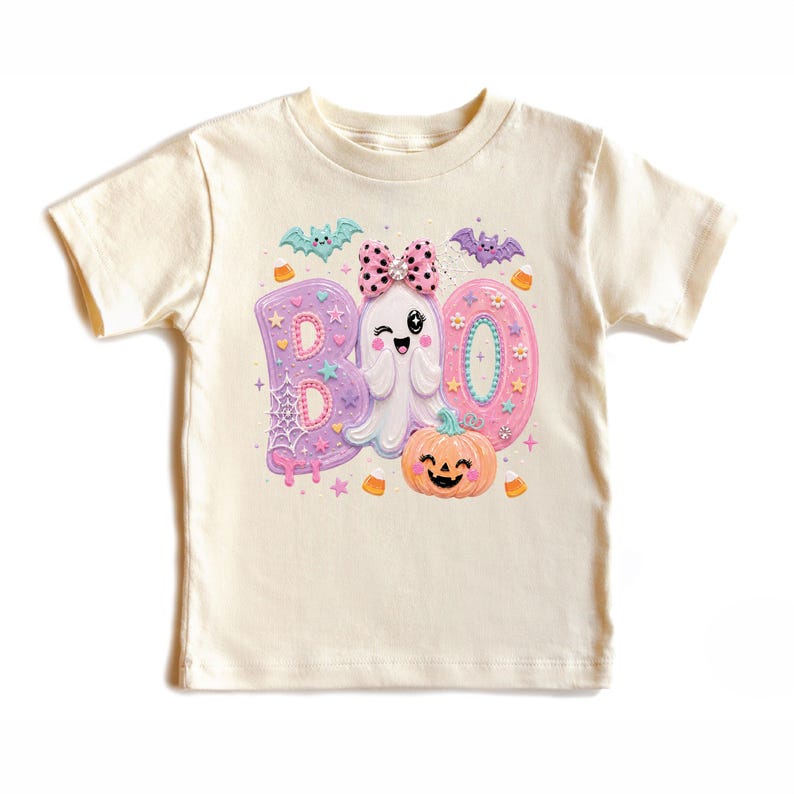 Puede incluir: Una camiseta de manga corta color crema con un estampado gr&aacute;fico de la palabra BOO en colores pastel. El dise&ntilde;o incluye un fantasma gui&ntilde;ando un ojo con un lazo rosa de lunares, una calabaza naranja sonriente, murci&eacute;lagos y telara&ntilde;as.
