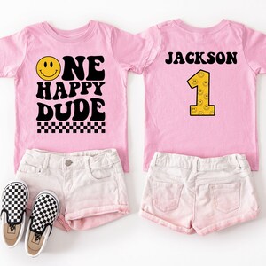 May include: Two pink t-shirts with the text "ONE HAPPY DUDE" and "JACKSON 1" in black and yellow. The shirts are paired with white shorts and black and white checkered sneakers. A fun outfit for a first birthday.