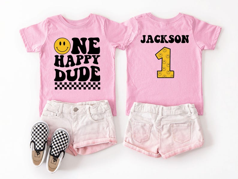 May include: Two pink t-shirts with the text "ONE HAPPY DUDE" and "JACKSON 1" in black and yellow. The shirts are paired with white shorts and black and white checkered sneakers. A fun outfit for a first birthday.