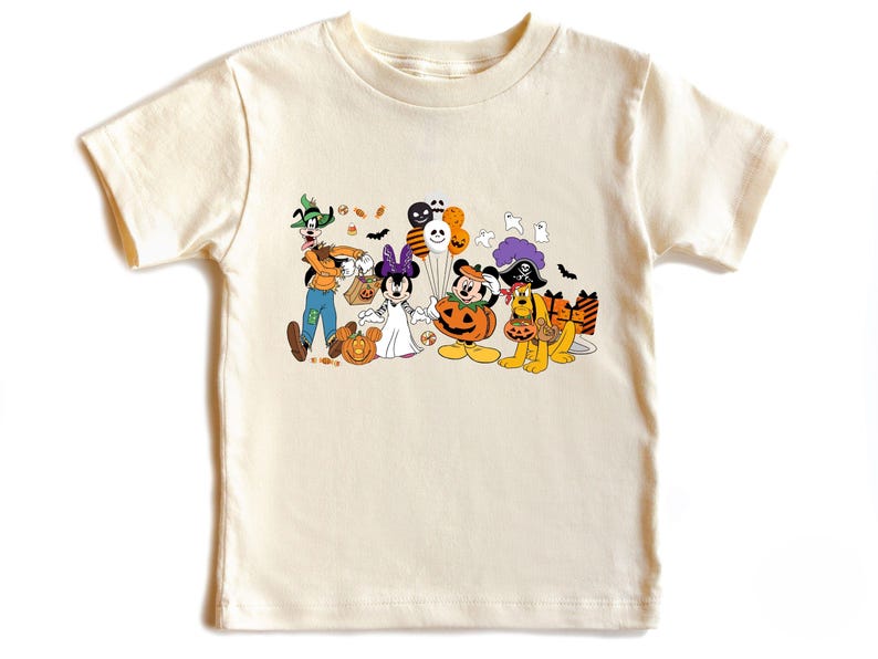 May include: Cream-colored t-shirt featuring a Halloween-themed graphic. The graphic includes cartoon characters dressed in costumes, such as a witch, a vampire, and a pirate. Pumpkins, bats, and balloons are also included in the design.