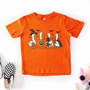 May include: Orange short-sleeved t-shirt featuring a Halloween-themed graphic of four cartoon geese in costumes. One goose wears a witch hat, another a pumpkin head, a third holds a black cat, and the last wears a cape and holds a coffee cup.