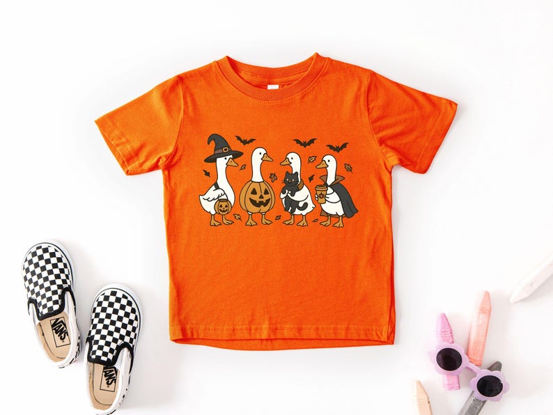 May include: Orange short-sleeved t-shirt featuring a Halloween-themed graphic of four cartoon geese in costumes. One goose wears a witch hat, another a pumpkin head, a third holds a black cat, and the last wears a cape and holds a coffee cup.