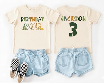 Könnte beinhalten: Zwei cremefarbene T-Shirts, eines mit dem Text "BIRTHDAY BOY" und Dinosaurier-Grafiken, das andere mit "JACKSON" und der Zahl "3". Dazu hellblaue Jeansshorts und ein Paar schwarz-weiß karierte Slip-on-Sneaker.