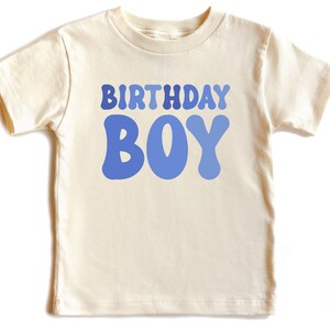 May include: A cream-coloured short-sleeved t-shirt featuring the words "BIRTHDAY BOY" in a blue, retro font. The shirt is made of a soft material, ideal for a birthday celebration.