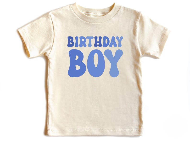May include: A cream-coloured short-sleeved t-shirt featuring the words "BIRTHDAY BOY" in a blue, retro font. The shirt is made of a soft material, ideal for a birthday celebration.