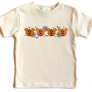 May include: Cream-colored t-shirt featuring a Halloween design. The design includes four orange pumpkin faces with mouse ears, black bats, and spiderwebs. The shirt is made of a soft material and is suitable for children.