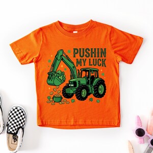 May include: Orange t-shirt with a green tractor graphic, a bucket of shamrocks, and the text "Pushin My Luck". The shirt is paired with black and white checkered slip-on shoes and sunglasses.