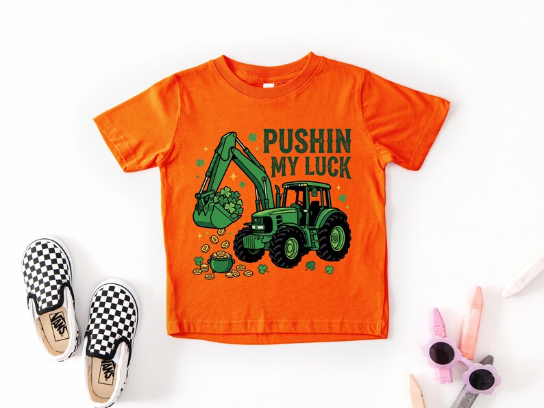 May include: Orange t-shirt with a green tractor graphic, a bucket of shamrocks, and the text "Pushin My Luck". The shirt is paired with black and white checkered slip-on shoes and sunglasses.