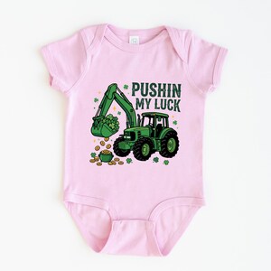 May include: Pink baby bodysuit with a green tractor graphic. The tractor's bucket is filled with shamrocks, and the text "PUSHIN MY LUCK" is above the tractor. Gold coins and shamrocks are scattered around the tractor.