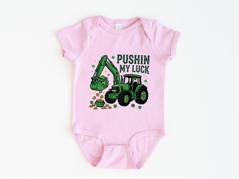 May include: Pink baby bodysuit with a green tractor graphic. The tractor's bucket is filled with shamrocks, and the text "PUSHIN MY LUCK" is above the tractor. Gold coins and shamrocks are scattered around the tractor.