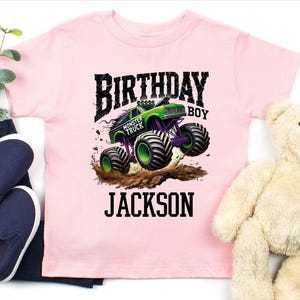 Pode incluir: Camiseta rosa claro com um gr&aacute;fico de monster truck verde e roxo. As palavras "BIRTHDAY BOY" e "JACKSON" s&atilde;o impressas em preto. Sapatos slip-on azul marinho e um ursinho de pel&uacute;cia tamb&eacute;m s&atilde;o vis&iacute;veis.
