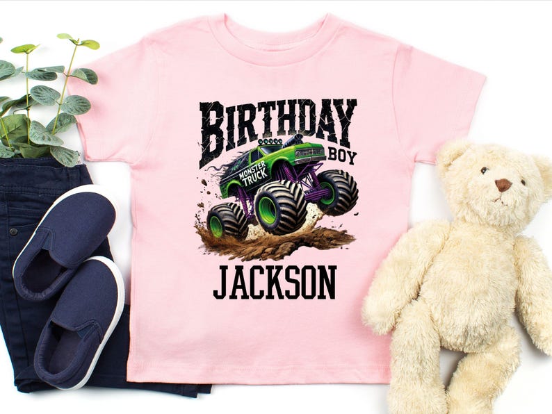 Pode incluir: Camiseta rosa claro com um gr&aacute;fico de monster truck verde e roxo. As palavras "BIRTHDAY BOY" e "JACKSON" s&atilde;o impressas em preto. Sapatos slip-on azul marinho e um ursinho de pel&uacute;cia tamb&eacute;m s&atilde;o vis&iacute;veis.