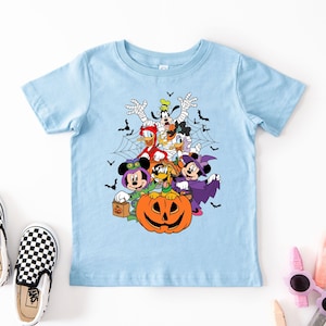 Op de afbeelding: Een lichtblauw T-shirt met een Halloween-ontwerp met Disney-personages in kostuums. Mickey en Minnie Mouse, Donald en Daisy Duck en Goofy worden afgebeeld. Er is ook een pompoen. Zwart-wit geblokte instappers liggen naast het shirt.