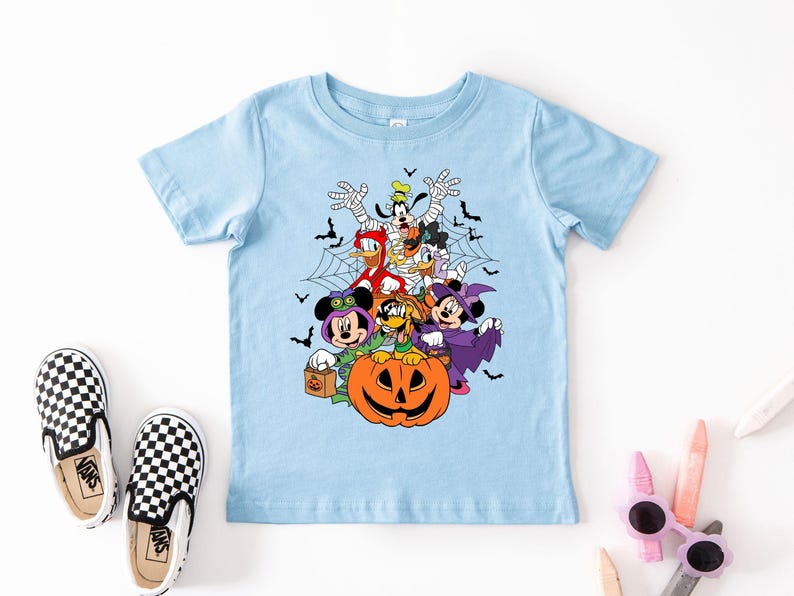 Op de afbeelding: Een lichtblauw T-shirt met een Halloween-ontwerp met Disney-personages in kostuums. Mickey en Minnie Mouse, Donald en Daisy Duck en Goofy worden afgebeeld. Er is ook een pompoen. Zwart-wit geblokte instappers liggen naast het shirt.
