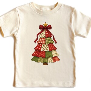 May include: Cream-colored t-shirt featuring a Christmas tree design. The tree is composed of red, green, and cream-colored patchwork patterns, topped with a gold star and a red bow. The tree has a brown trunk.