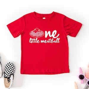 May include: A red t-shirt with the words "one little meatball" in white. The design includes a spaghetti and meatball graphic. Black and white checkered slip-on shoes and sunglasses are also in the image.