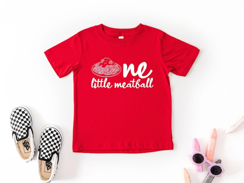 May include: A red t-shirt with the words "one little meatball" in white. The design includes a spaghetti and meatball graphic. Black and white checkered slip-on shoes and sunglasses are also in the image.