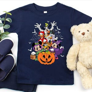 Op de afbeelding: Marineblauwe peuter-t-shirt met een Halloween-thema met Disney-personages, waaronder Mickey en Minnie, Donald Duck en Goofy, rond een uitgesneden pompoen. Het ontwerp bevat vleermuizen en spinnenwebben. Het shirt wordt gecombineerd met een marineblauwe broek en schoenen.