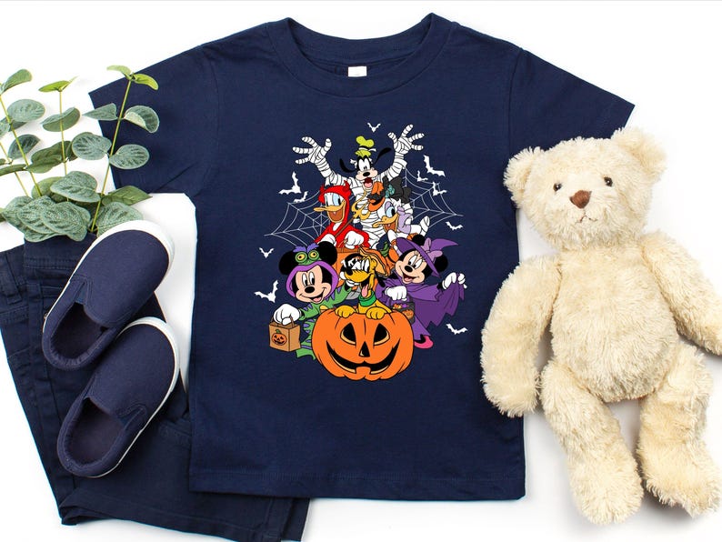 Op de afbeelding: Marineblauwe peuter-t-shirt met een Halloween-thema met Disney-personages, waaronder Mickey en Minnie, Donald Duck en Goofy, rond een uitgesneden pompoen. Het ontwerp bevat vleermuizen en spinnenwebben. Het shirt wordt gecombineerd met een marineblauwe broek en schoenen.