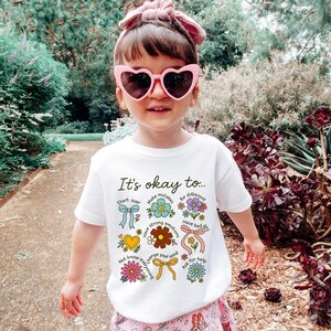 May include: A young person wearing a white t-shirt with the text "It's okay to..." and various floral and ribbon graphics. The person is also wearing pink heart-shaped sunglasses and a pink headband. The background features lush greenery.