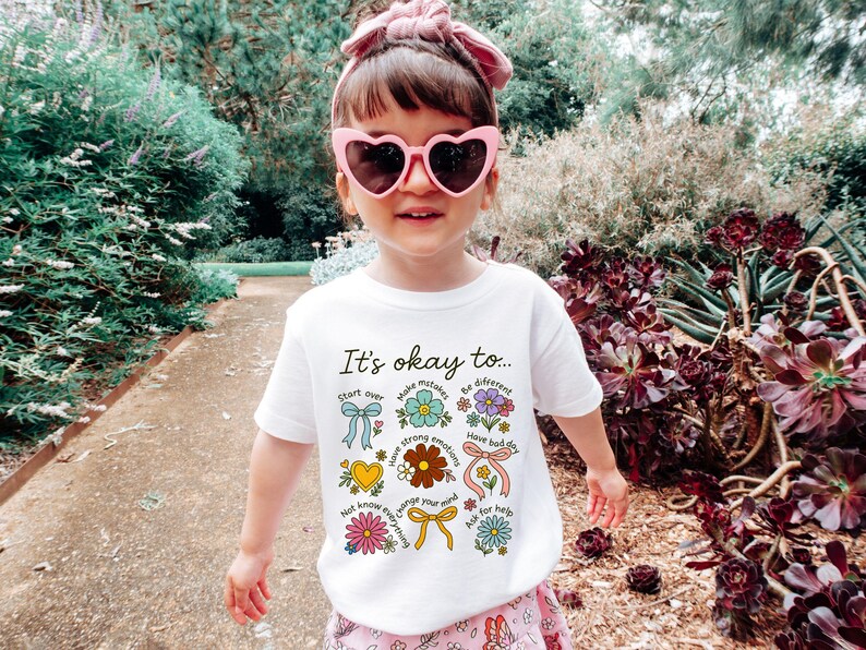 May include: A young person wearing a white t-shirt with the text "It's okay to..." and various floral and ribbon graphics. The person is also wearing pink heart-shaped sunglasses and a pink headband. The background features lush greenery.