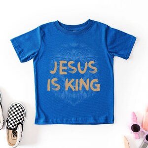 May include: A blue t-shirt with the text "JESUS IS KING" in gold lettering. A lion's face is subtly visible behind the text. Checkered black and white slip-on shoes and sunglasses are also in the image.