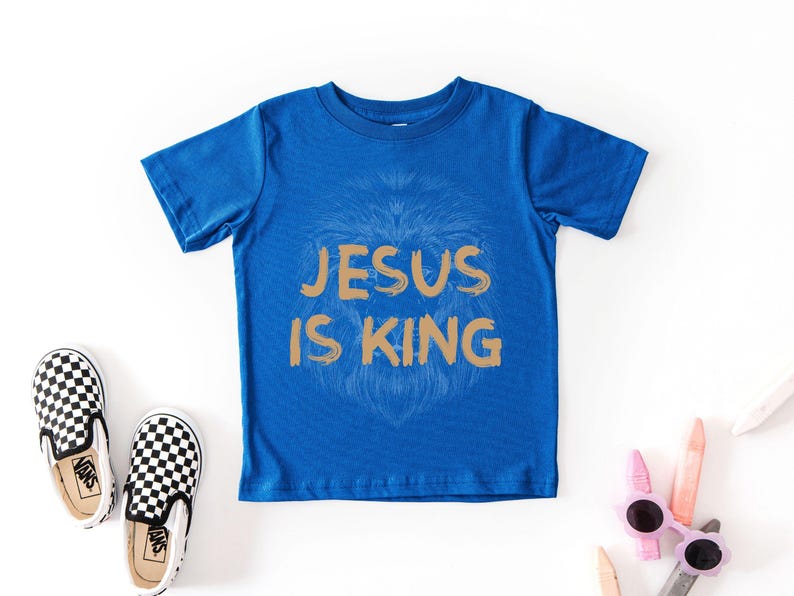 May include: A blue t-shirt with the text "JESUS IS KING" in gold lettering. A lion's face is subtly visible behind the text. Checkered black and white slip-on shoes and sunglasses are also in the image.