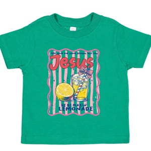 May include: A green toddler t-shirt with a graphic design. The design features the word "Jesus" in pink, a glass of lemonade, and a lemon slice. The text "Life gave me lemons but God" and "He be makin lemonade" are also included.