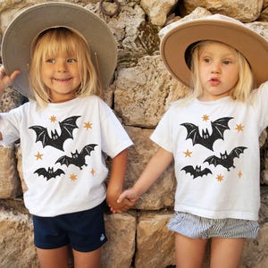 May include: Two white short-sleeved t-shirts featuring a black graphic print of three bats and several small orange stars. The shirts are worn with a grey wide-brimmed hat, navy blue shorts, and grey and white striped shorts respectively.
