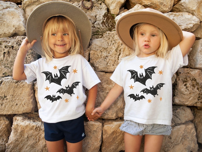 May include: Two white short-sleeved t-shirts featuring a black graphic print of three bats and several small orange stars. The shirts are worn with a grey wide-brimmed hat, navy blue shorts, and grey and white striped shorts respectively.