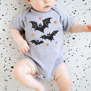 May include: A heather gray short-sleeve baby bodysuit featuring a graphic print of three black bats and four small orange stars. The garment has a round neckline and a snap-button closure at the bottom, designed for infants with a simple, functional fit.