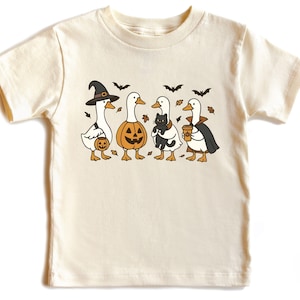 May include: Cream-colored t-shirt featuring a Halloween-themed graphic. The design includes four cartoon geese dressed in costumes: a witch, a jack-o'-lantern, a goose holding a black cat, and a vampire. Bats and leaves are also included.