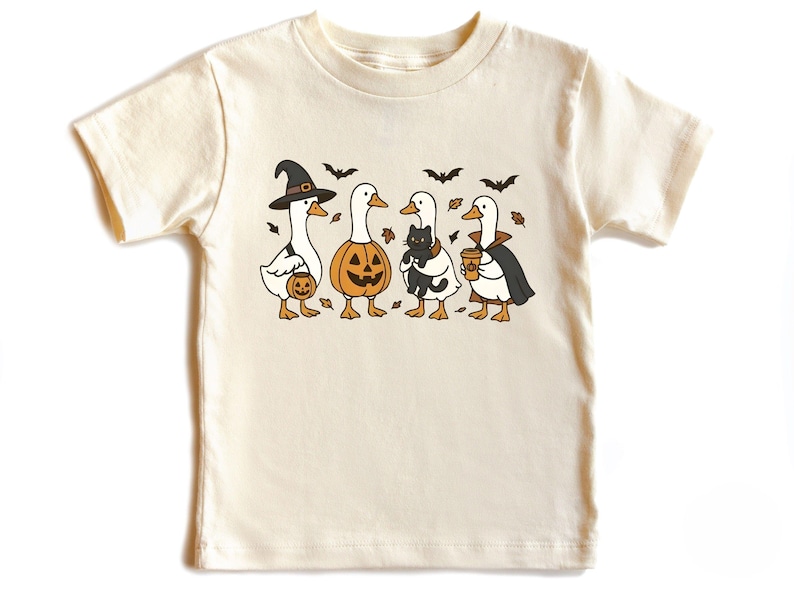 May include: Cream-colored t-shirt featuring a Halloween-themed graphic. The design includes four cartoon geese dressed in costumes: a witch, a jack-o'-lantern, a goose holding a black cat, and a vampire. Bats and leaves are also included.