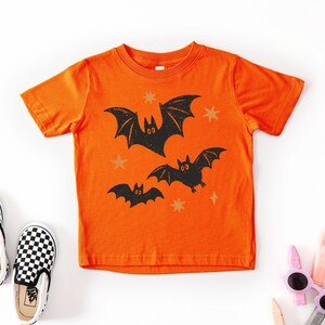 May include: A bright orange short-sleeve t-shirt featuring a graphic print of three black bats in flight, accented by small yellow star shapes scattered across the front. The design has a textured, distressed appearance on the fabric.