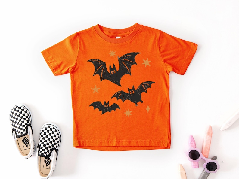 May include: A bright orange short-sleeve t-shirt featuring a graphic print of three black bats in flight, accented by small yellow star shapes scattered across the front. The design has a textured, distressed appearance on the fabric.