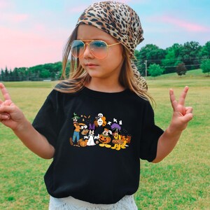 May include: Black t-shirt featuring a Halloween-themed graphic with cartoon characters, pumpkins, and ghosts. The person is wearing a leopard print scarf and sunglasses, posing outdoors with a peace sign.
