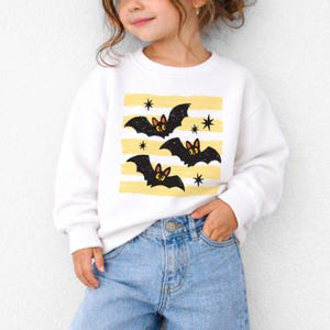 May include: A white long-sleeve sweatshirt featuring a graphic print of three black bats with orange ears and yellow eyes, set against horizontal yellow brushstroke stripes and small black star accents, paired with light blue denim jeans.