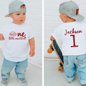 May include: A white t-shirt with the text "one little meatball" in red, worn by a toddler. The toddler is also wearing blue jeans and a gray baseball cap. The back of the shirt has the name "Jackson" and the number "1" in red.